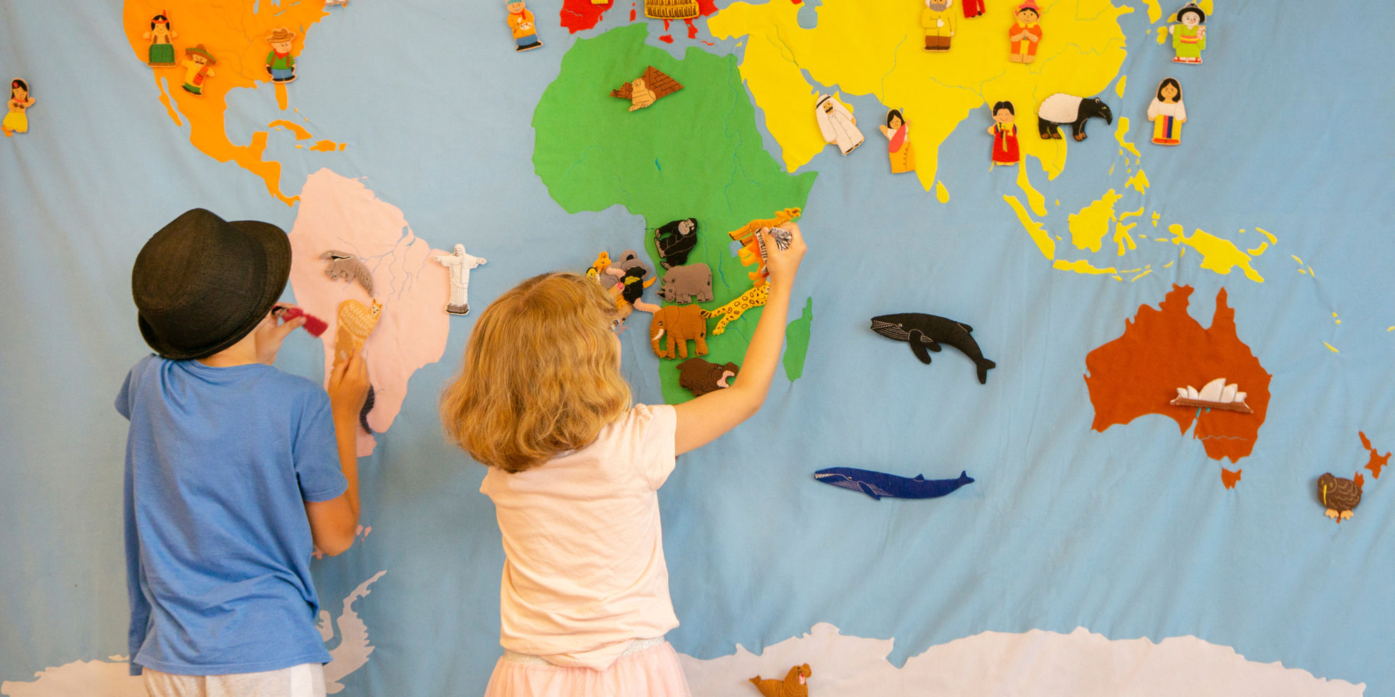 Zwei Kinder stehen vor einem großen Wandbild aus Stoff. Auf diesem sind Kontinente und Meere zu sehen. Sie pinnen bunte Tiere, Menschen und Gebäude aus Stoff auf das Wandbild.
