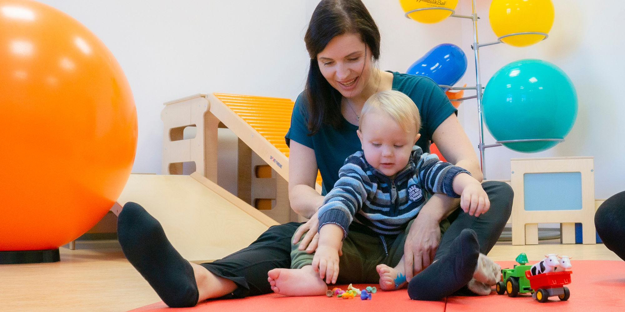© Adobe Stock: Köpenicker, #32724599 Eine Physiotherapeutin sitzt mit einem kleinen Jungen auf einer Turnmatte und positioniert seine Füße während er versucht nach bunten Figuren zu greifen.
