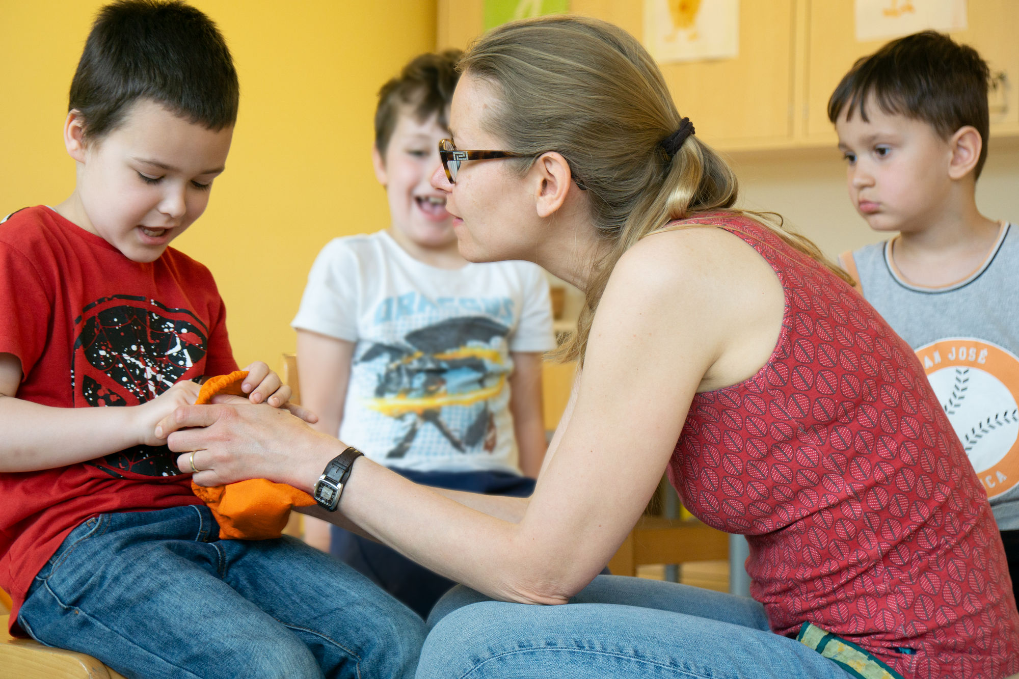 Eine Frau hält einen kleinen orangefarbenen Sack zu fühlen einem Jungen hin. Im Hintergrund sind zwei weitere Kinder zu sehen