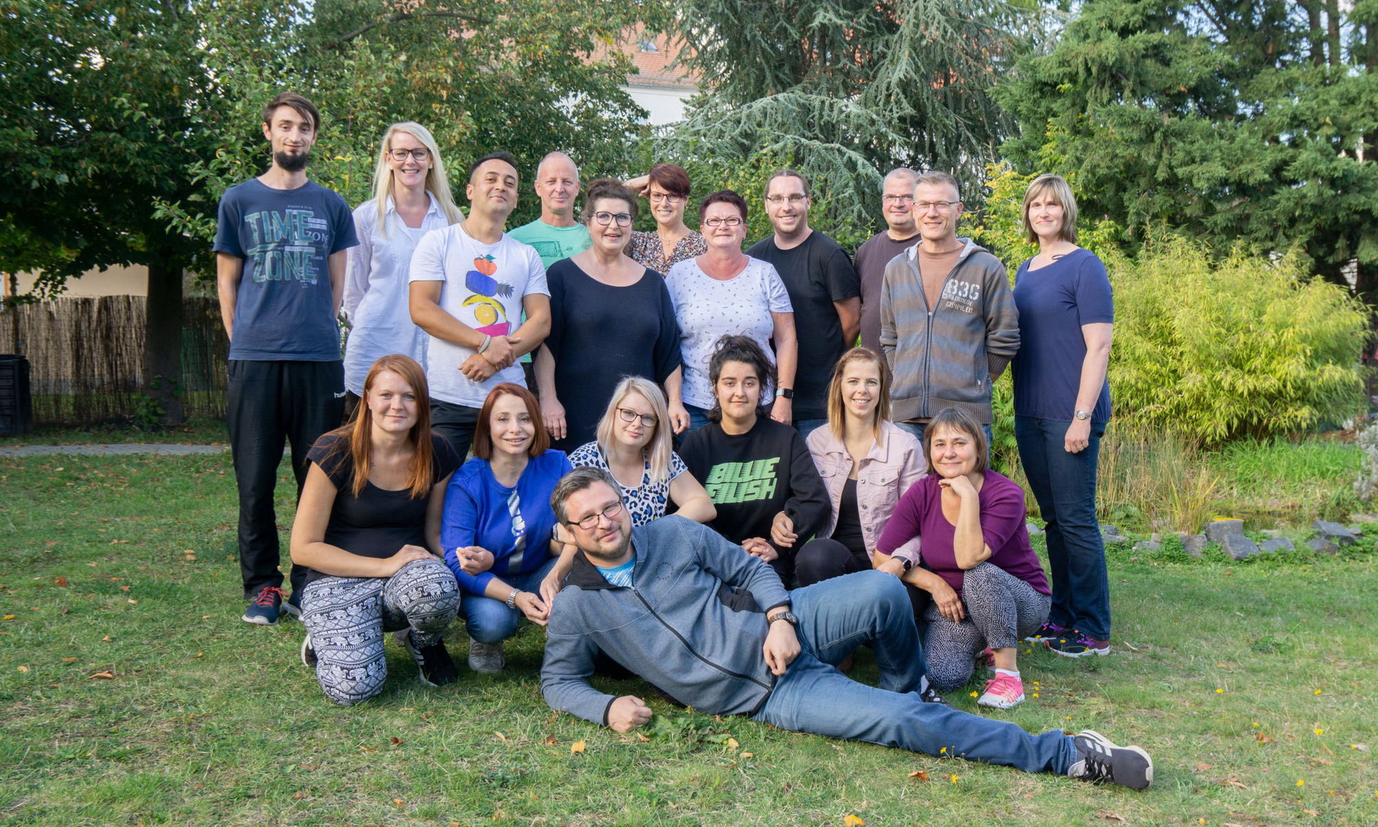 18 Mitarbeiter:innen befinden sich auf einer Wiese im Garten und lächeln in die Kamera.