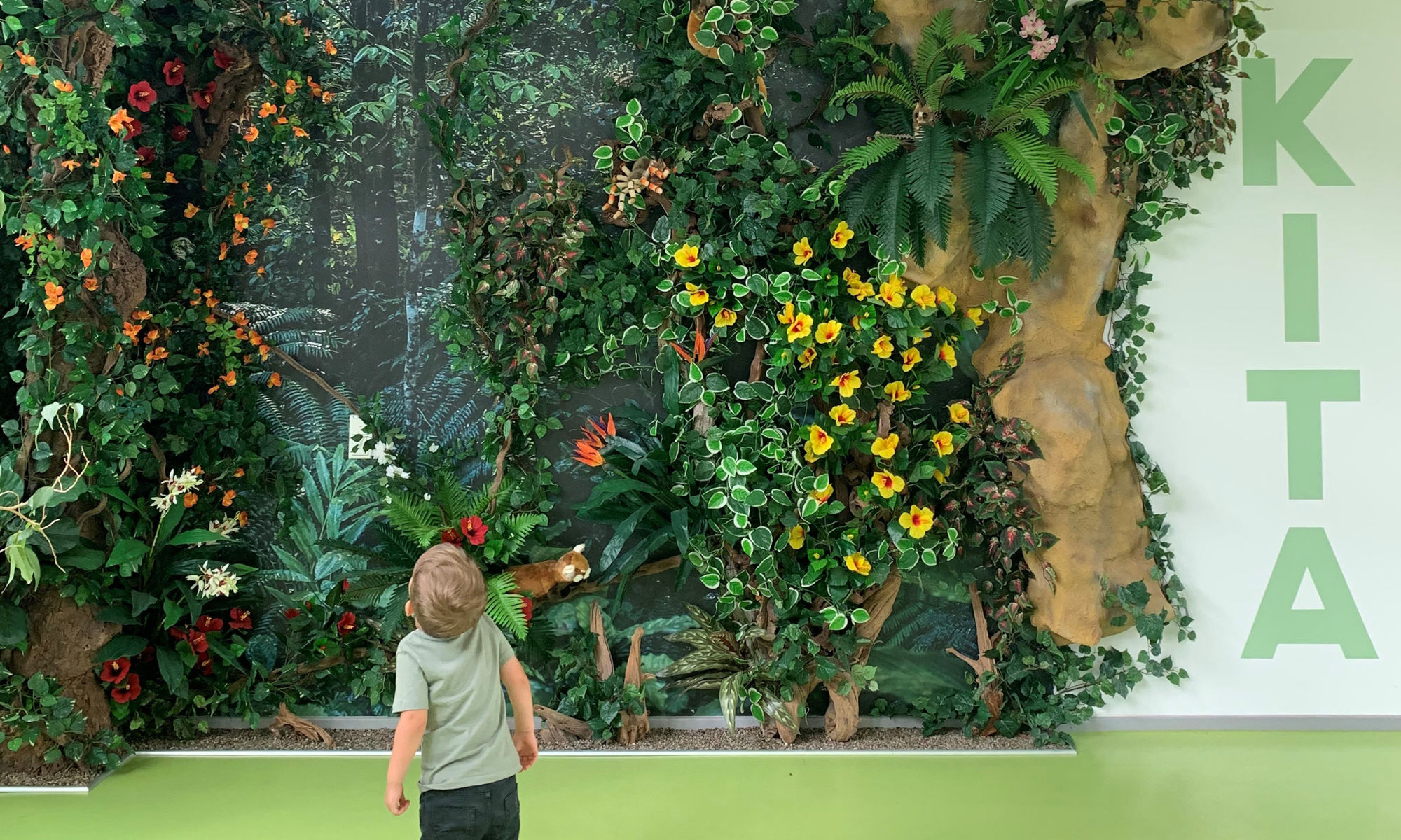 Ein Kind steht vor einer großen Wand mit Pflanzen, Lianen, kleinen Tieren und einem großen Baum.