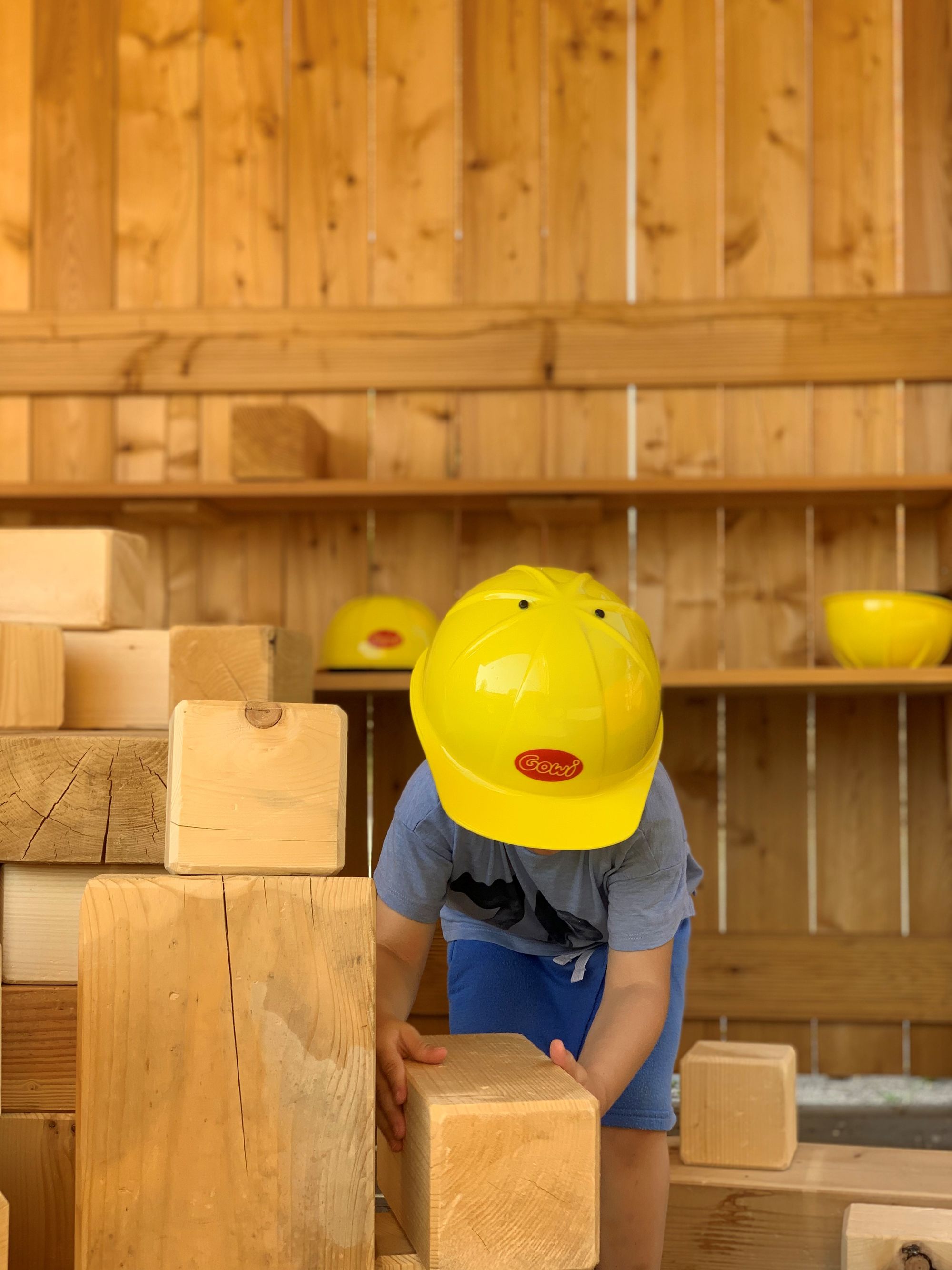Ein Junge stapelt große Holzblöcke und hat einen Bauhelm auf.