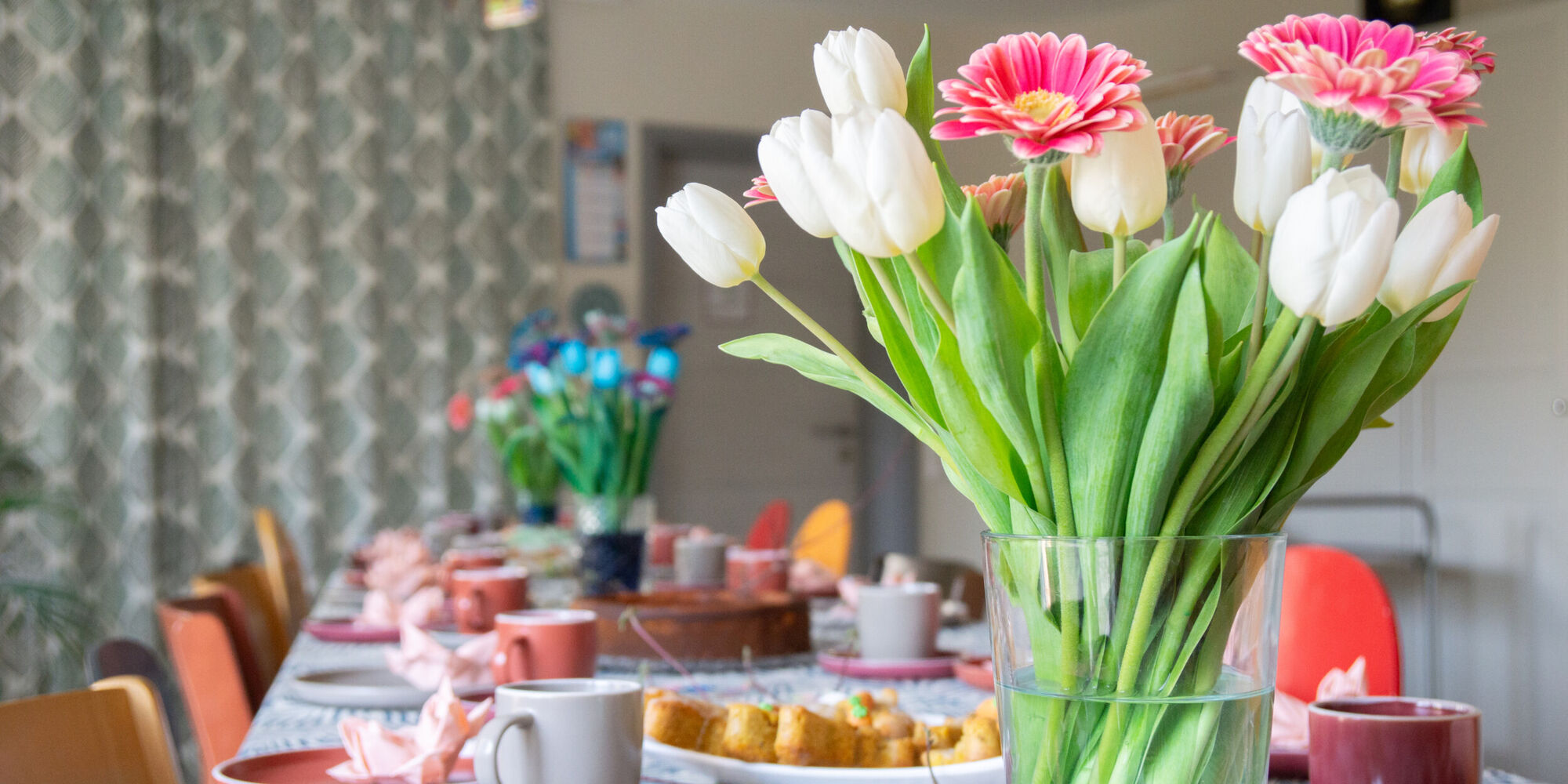 Ein Blumenstrauß mit weißen Tulpen und pinkfarbenen Gerberas steht auf einer gedeckten Kaffeetafel mit Kuchen.