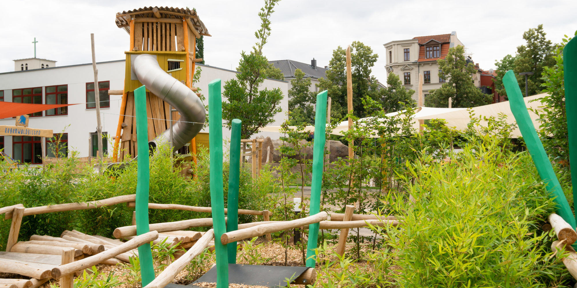 Ein großer Holzturm mit Röhrenrutsche, davor viele grüne Pflanzen und ein Kletterparcours.