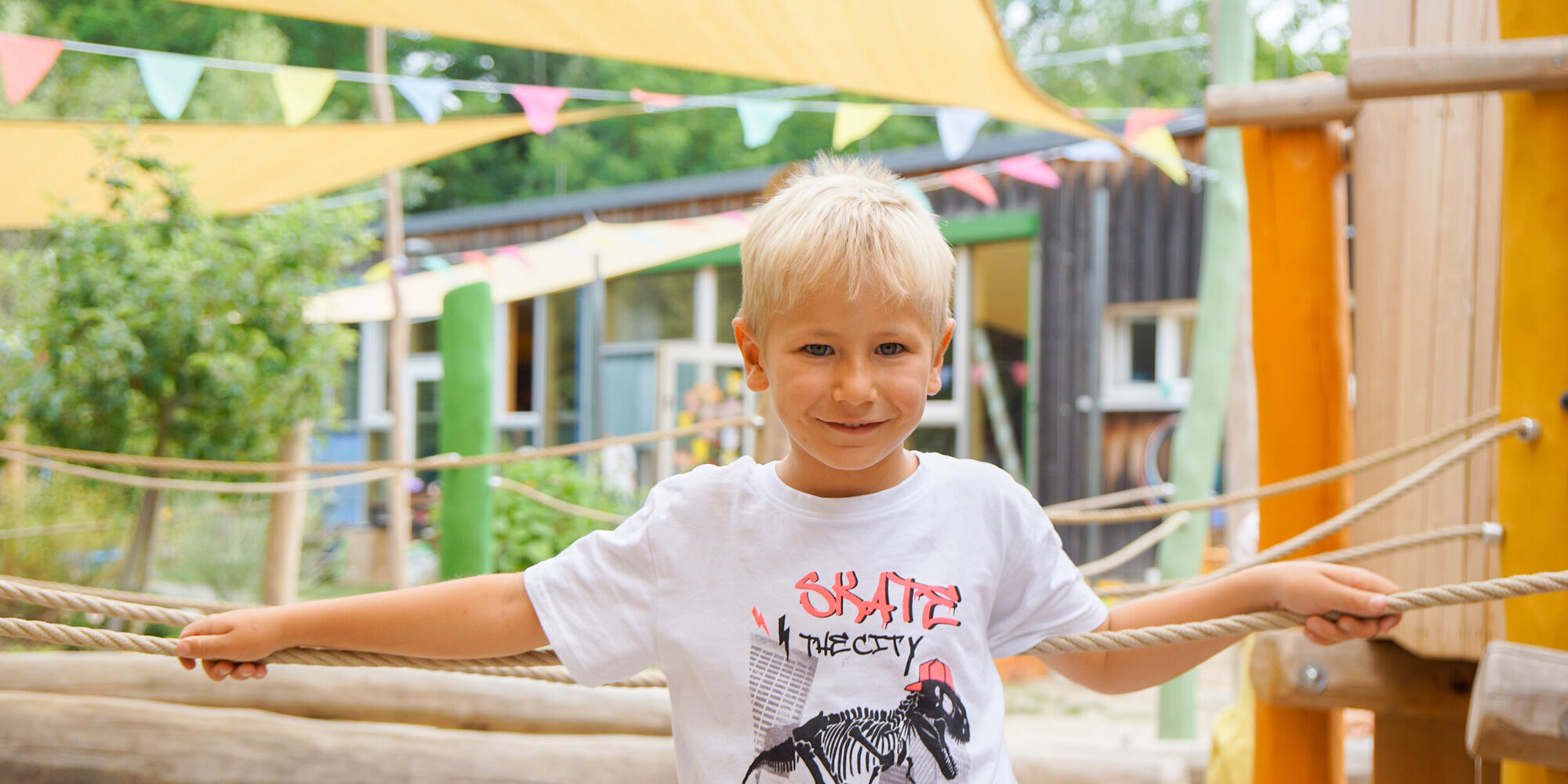 Ein Junge steht auf einem Kletterparcours und hält sich an Seilen fest. Er lacht.