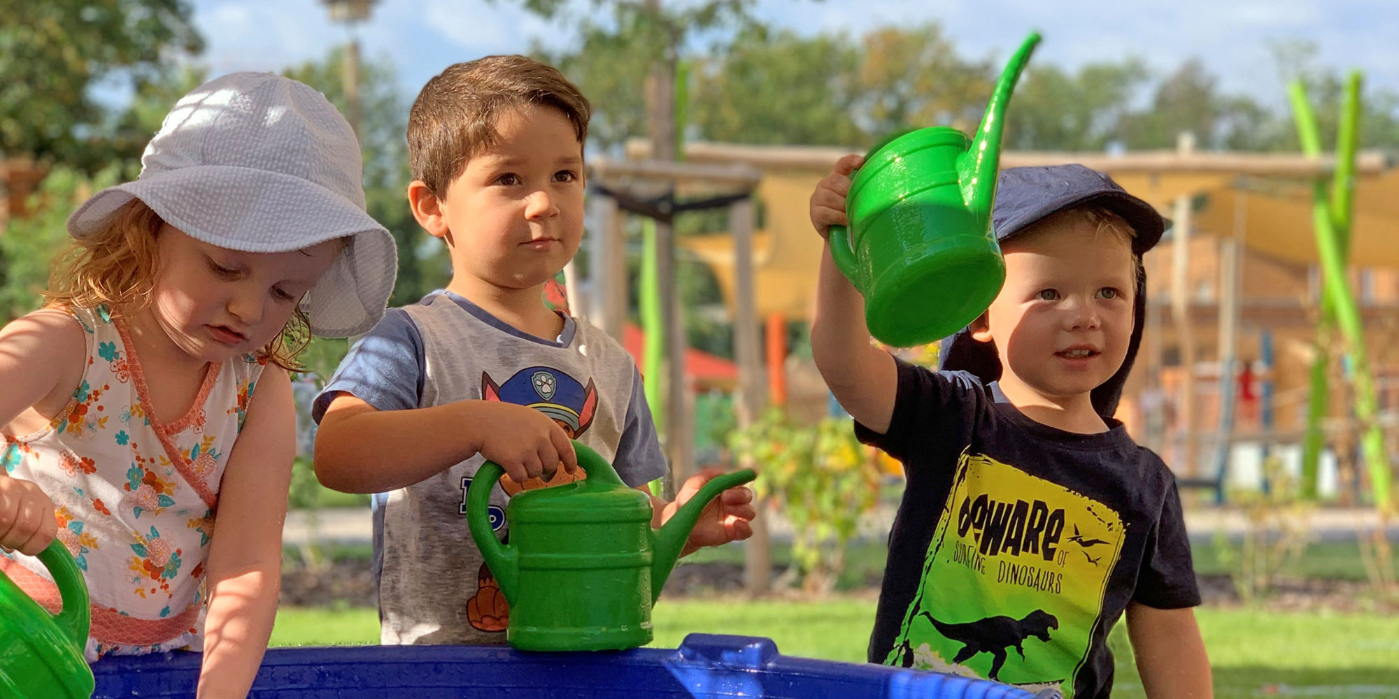 Drei Kleine Kinder stehen an einem blauen Wasserbecken und füllen Gießkannen auf.