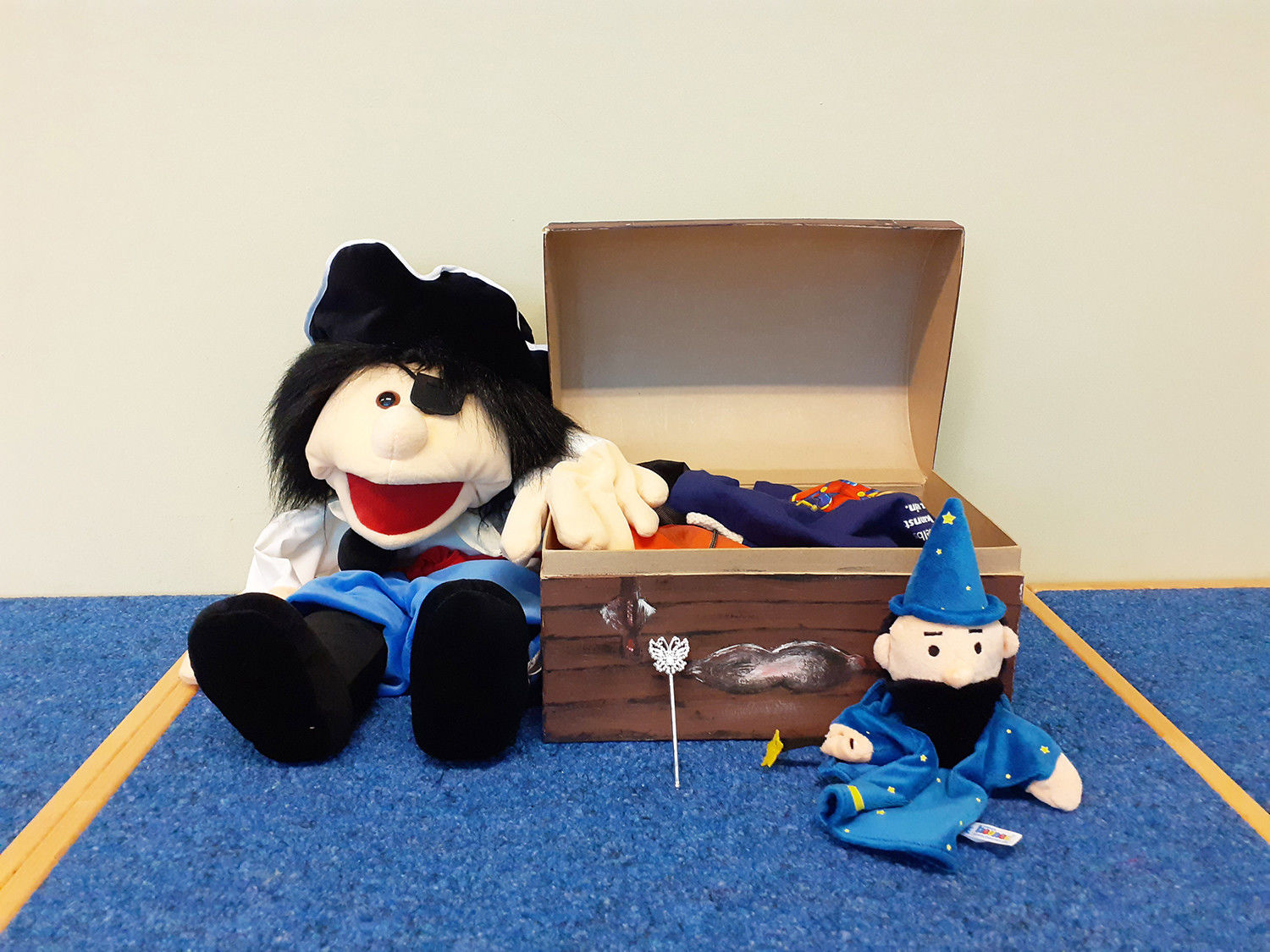 Ein Piratenpuppe aus Plüsch hat den Arm auf eine geöffnete Schatztruhe gelegt. Davor sitzt eine Handpuppe mit Zauberstab und spitzem blauen Hut.