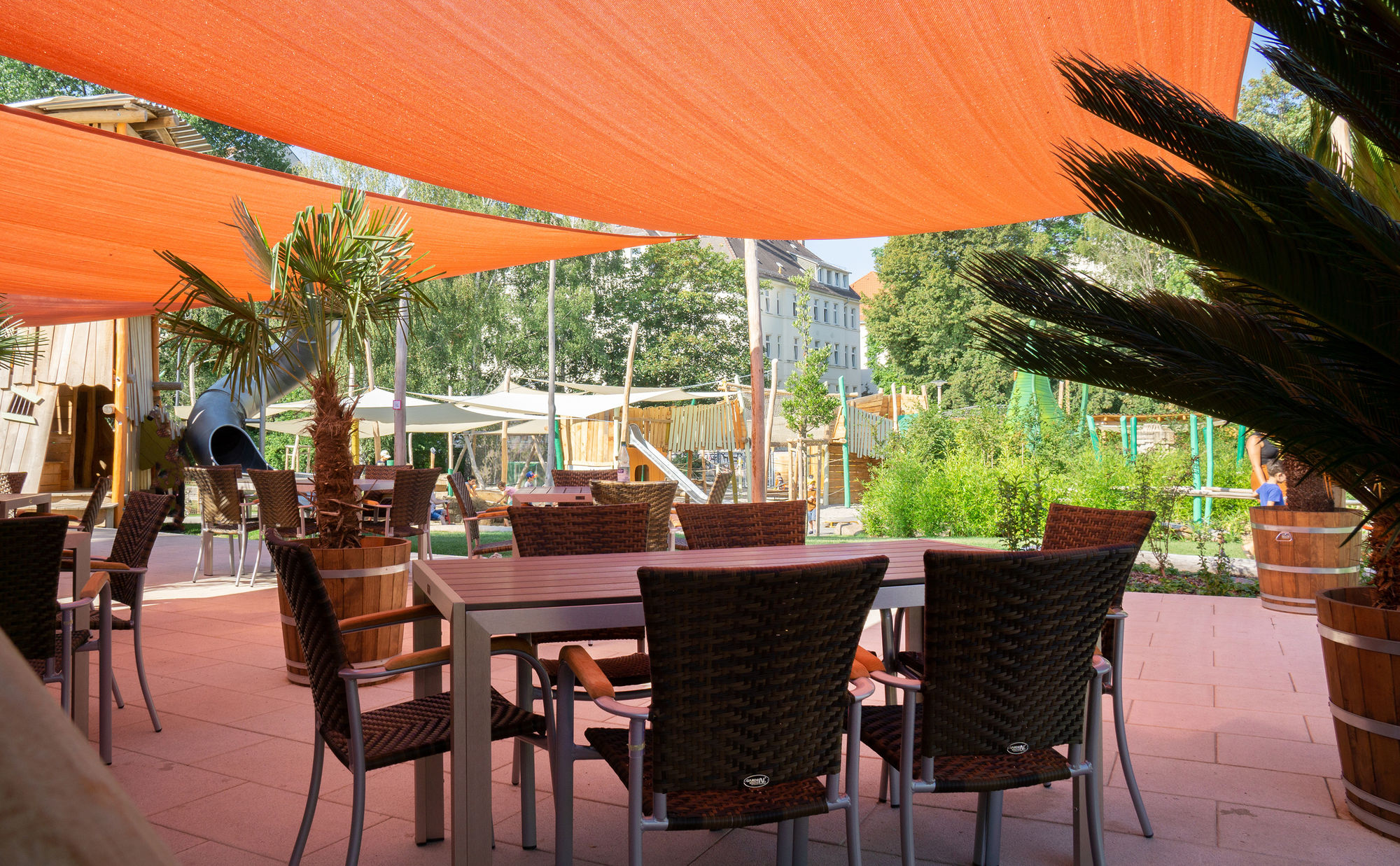 Ein mit rotem Sonnensegel überdachte Terrasse mit Tischen, Stühlen und Palmen.
