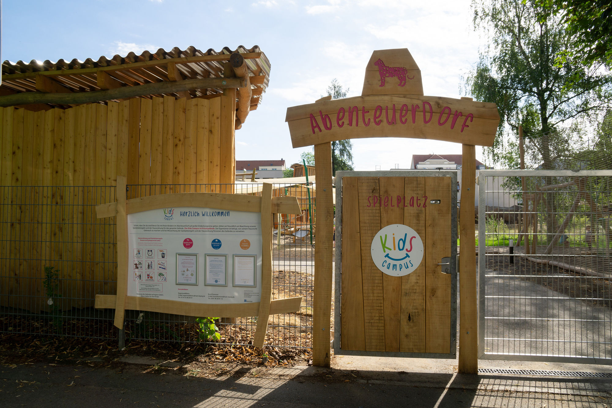 Ein helles Holztor in einem Gitterzaun. Darauf ist ein weißer Kreis mit dem bunten Kids Campus Logo zu sehen. darüber ein geschwungenes Brett mit pinker Aufschrift AbenteuerDorf und einem Geparden.
