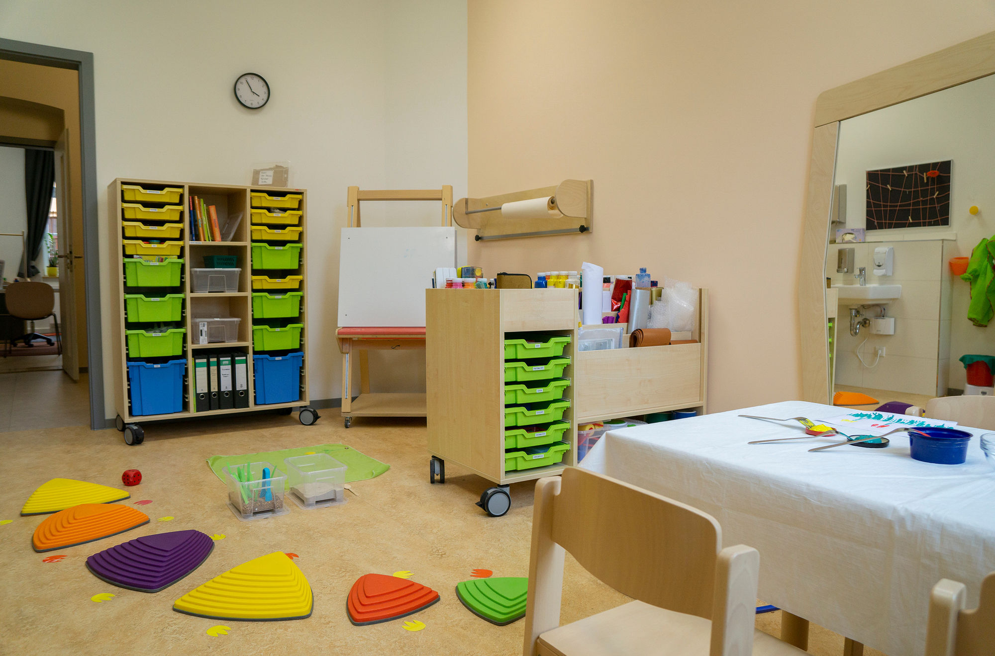 Ein heller Raum mit Regalen mit bunten Kisten, einer Leinwand, einem Tisch, Stühlen und bunten Trittsteinen auf dem Boden.