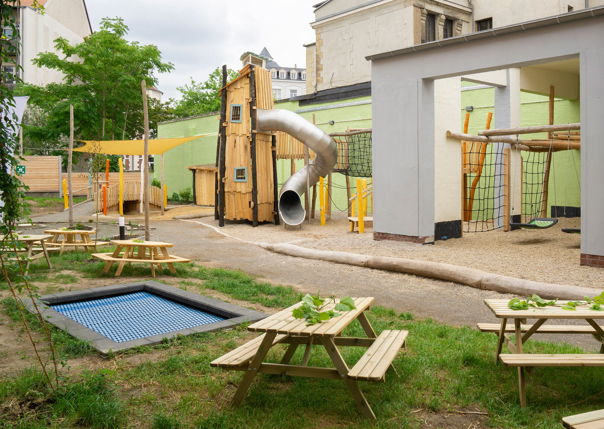 Ein großer Rutschenturm, Klettermöglichkeiten, ein Trampolin und kleine Sitzbänke mit Tischen.
