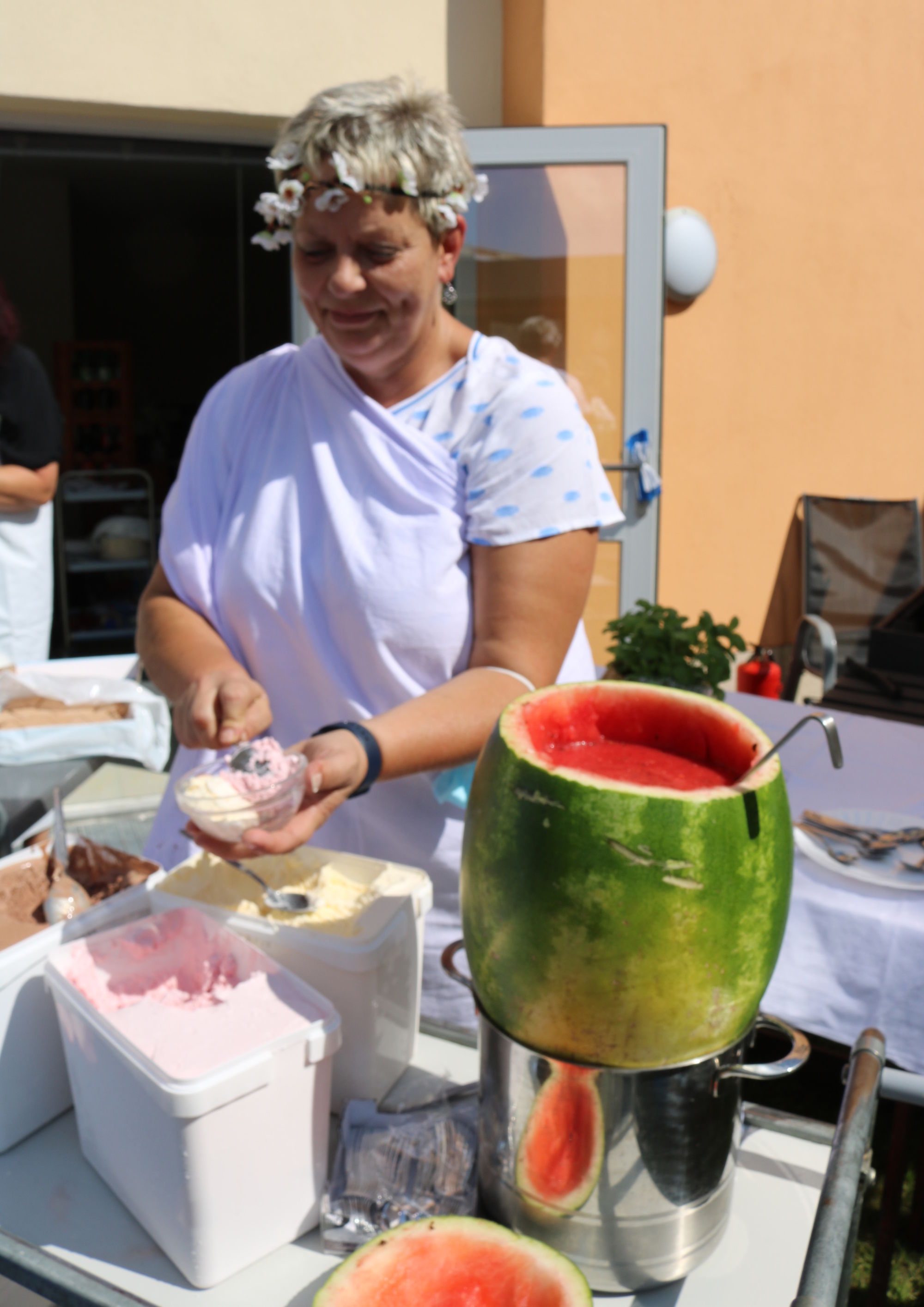 Eine Frau mit Blumenkranz auf dem Kopf steht hinter einem Tisch mit einer Melonenbowle und Eiscreme und tut Eiskugeln in eine kleine Schüssel