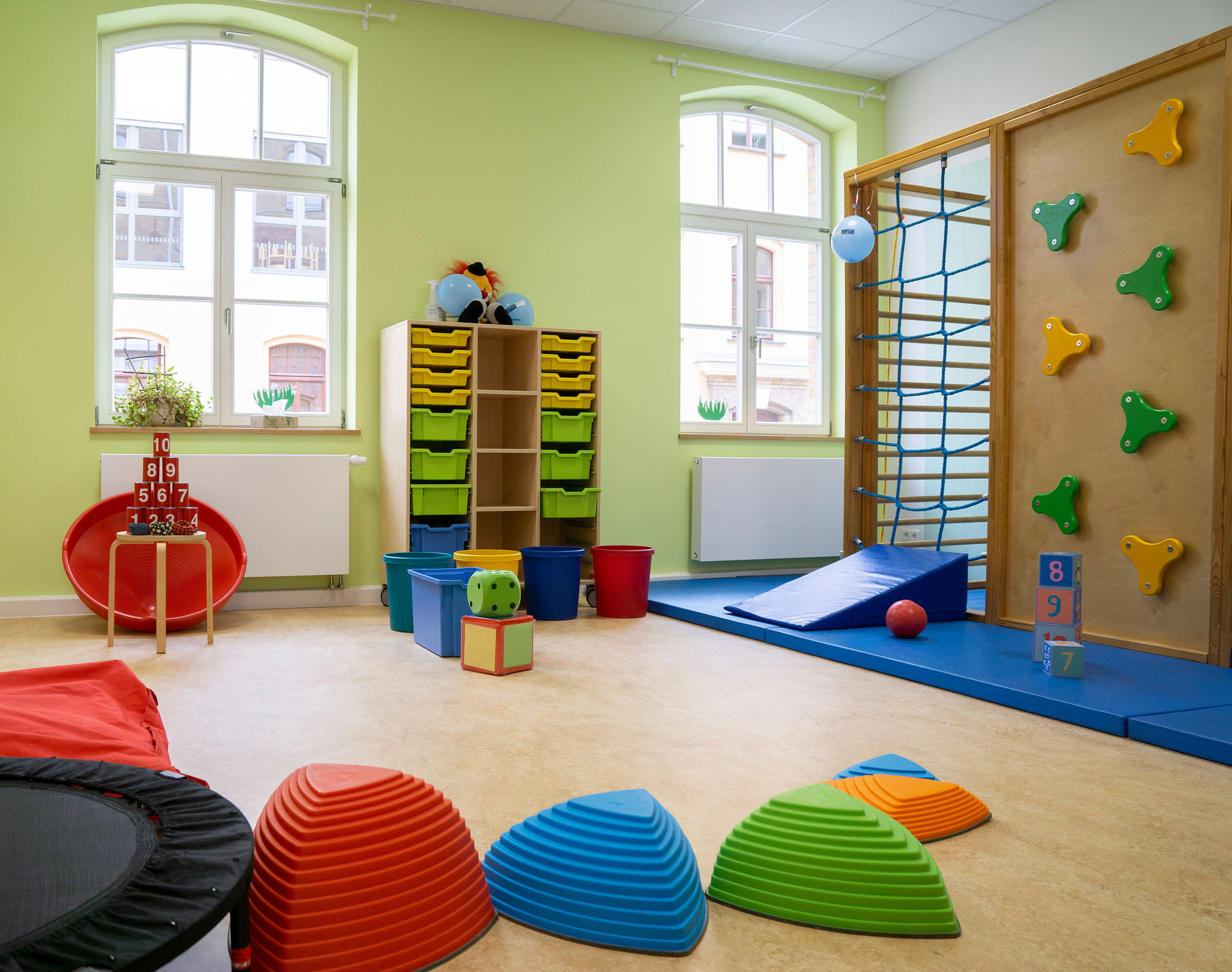 Ein heller Raum mit Sportmatten, Kletterwand, Trampolin und bunten Trittsteinen.
